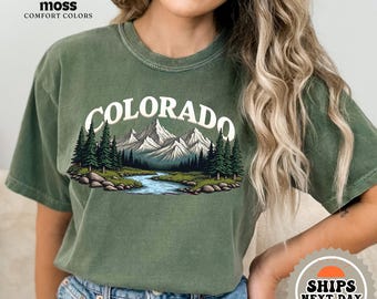 Puede incluir: Una camiseta verde con un gráfico de montañas y árboles. El texto "COLORADO" está impreso en blanco en la camiseta.
