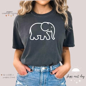May include: A dark gray t-shirt with a white outline of an elephant. The text "pepper COMFORT COLORS" is at the top of the shirt. The text "also available in sweatshirt & hoodie" is at the bottom of the shirt.