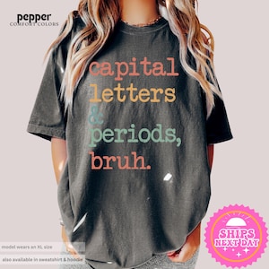 K&ouml;nnte beinhalten: Ein dunkelgraues T-Shirt mit dem Text "capital letters & periods, bruh." in bunten Buchstaben.