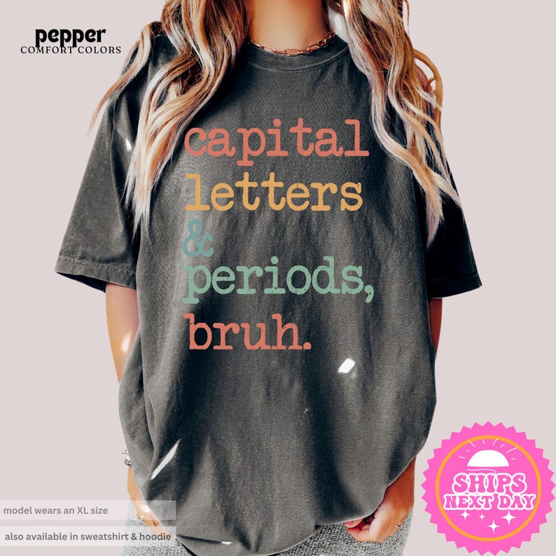 K&ouml;nnte beinhalten: Ein dunkelgraues T-Shirt mit dem Text "capital letters & periods, bruh." in bunten Buchstaben.