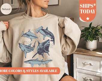 May include: A beige sweatshirt with a graphic print of various sharks and a hammerhead shark. The text "MORE COLORS & STYLES AVAILABLE" is printed on the bottom of the sweatshirt.