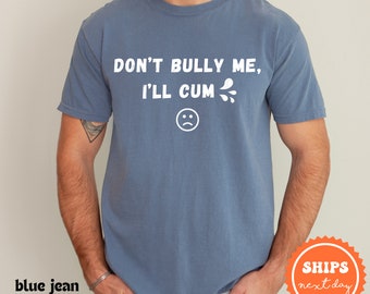 以下が含まれることがあります： 白い文字で「DON'T BULLY ME, I'LL CUM」と書かれた青いTシャツ。テキストの下には白い悲しい顔の絵文字があります。