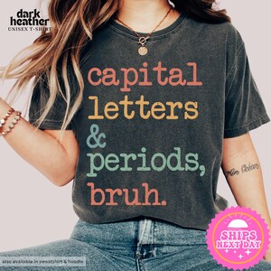 K&ouml;nnte beinhalten: Ein dunkel-heides graues T-Shirt mit dem Text "capital letters & periods, bruh." in bunten Buchstaben.