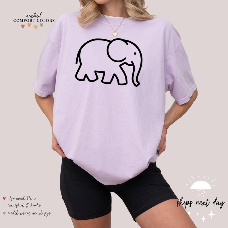 May include: A lavender t-shirt with a black outline of an elephant. The text "orchid COMFORT COLORS" is at the top of the shirt. The model is wearing the shirt with black biker shorts.