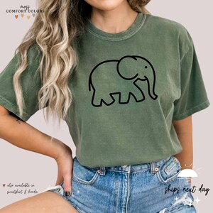 May include: A green t-shirt with a black outline of an elephant. The text "mozi COMFORT COLORS" is at the top of the image. The text "also available in sweatshirt & hoodie" is at the bottom of the image.
