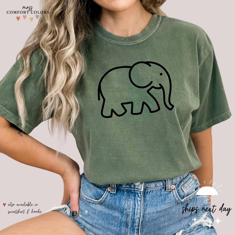 May include: A green t-shirt with a black outline of an elephant. The text "mozi COMFORT COLORS" is at the top of the image. The text "also available in sweatshirt & hoodie" is at the bottom of the image.