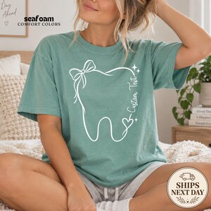 May include: A seafoam green short-sleeve t-shirt featuring a white line art graphic of a tooth adorned with a bow and a heart. The text "Custom Text" is printed vertically along the right side of the tooth design, accompanied by small star accents.