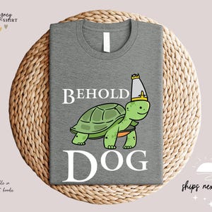 May include: A grey t-shirt featuring a cartoon turtle wearing a crown and the text "BEHOLD DOG" printed on it.