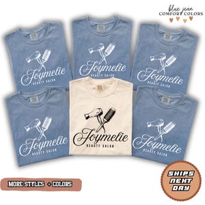 May include: A collection of blue and cream-colored t-shirts. Each shirt features a graphic of a hairdryer and a brush, with the text "Joymelie BEAUTY SALON" in a script font. The shirts are displayed on a white background.