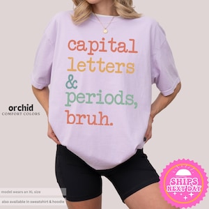 K&ouml;nnte beinhalten: Ein helles lila T-Shirt mit dem Text "capital letters & periods, bruh." in Regenbogenfarben.