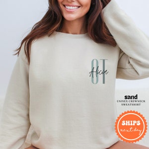 May include: A beige sweatshirt with the letters "OT" in a light blue and gray color scheme. The name "Alicia" is written in black script below the letters.