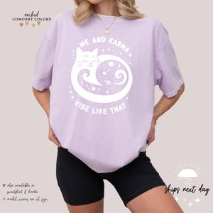 K&ouml;nnte beinhalten: Ein lavendelfarbenes T-Shirt mit einer wei&szlig;en Grafik eines Katers, der sich in einem Kreis mit Sternen und Planeten darin zusammengerollt hat. Der Text "Me and Karma Vibe Like That" ist unter dem Kater aufgedruckt.
