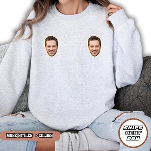 May include: A light gray crewneck sweatshirt featuring two identical photographic prints of a smiling man's face positioned on the upper chest area. The garment has long sleeves and a ribbed neckline, cuffs, and hem, providing a standard fit.