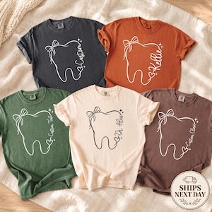 May include: Five short-sleeved t-shirts in charcoal, burnt orange, olive green, cream, and brown. Each shirt features a white or black line-art graphic of a tooth with a bow, sparkles, a heart, and custom text including Custom, Kellie, and Dr. Alex.