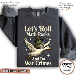 May include: A dark heather gray crewneck sweatshirt featuring a graphic of a frog in a wizard hat riding a flying white goose. The frog holds a twenty-sided die. The text reads: Let's Roll Math Rocks And Do War Crimes, surrounded by small stars.