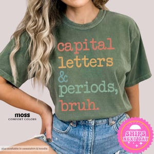 K&ouml;nnte beinhalten: Ein gr&uuml;nes T-Shirt mit dem Text "capital letters & periods, bruh." in bunten Buchstaben.