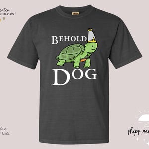 May include: Dark heather grey t-shirt featuring a cartoon image of a turtle wearing a crown and the text "Behold Dog".
