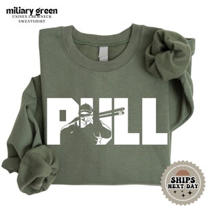 May include: A military green sweatshirt with the word "PULL" in white text. The word "PULL" is formed by a silhouette of a person holding a shotgun.