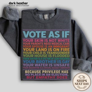 May include: A dark heather gray crewneck sweatshirt featuring multi-colored text that reads: VOTE AS IF YOUR SKIN IS NOT WHITE, YOUR PARENTS NEED MEDICAL CARE, YOUR SPOUSE IS AN IMMIGRANT, YOUR LAND IS ON FIRE, YOUR CHILD IS TRANSGENDER, YOUR HOUSE IS FLOODED, YOUR SISTER IS A VICTIM OF GUN VIOLENCE, YOUR BROTHER IS GAY, YOUR WATER IS UNSAFE, YOUR DAUGHTER IS A SEXUAL ASSAULT SURVIVOR, BECAUSE PRIVILEGE HAS NO PLACE IN AN ELECTION, BUT EMPATHY DOES.