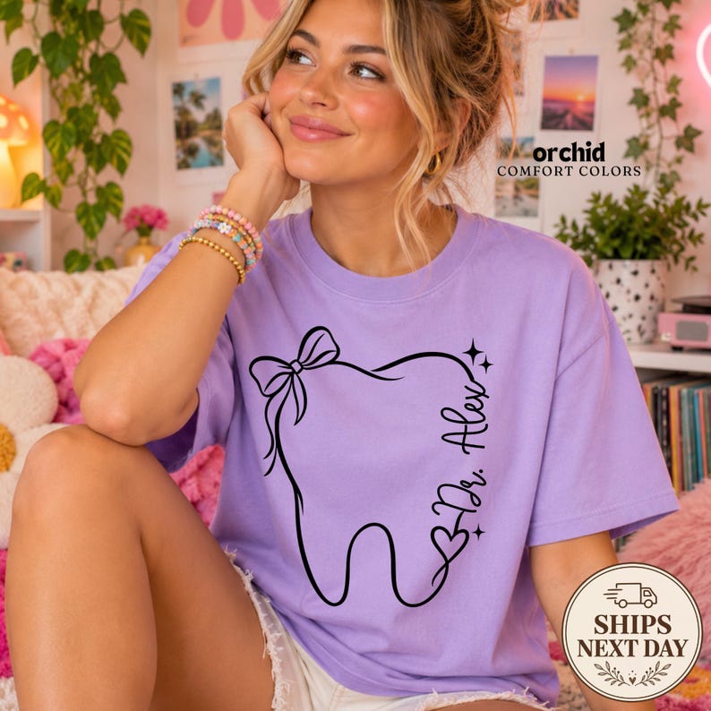 May include: A light purple short-sleeve t-shirt featuring a black line art graphic of a tooth with a bow on top and the text Dr. Alex written vertically along the side. The design includes small star accents and a heart shape near the bottom.