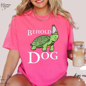May include: Pink t-shirt featuring a white graphic of a turtle wearing a crown and the text "Behold Dog".