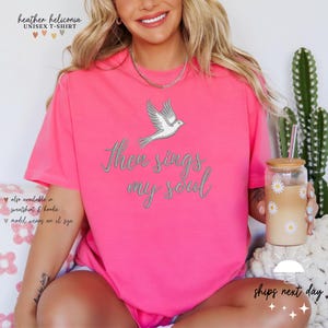 Puede incluir: Camiseta rosa con una paloma blanca y el texto "Then sings my soul" impreso en la parte delantera.