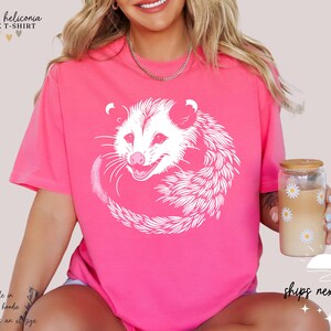 Puede incluir: Camiseta unisex rosa brillante con un dise&ntilde;o de zarig&uuml;eya blanca. La zarig&uuml;eya est&aacute; enroscada. El texto "heather heliconia UNISEX T-SHIRT" est&aacute; en la parte superior. Tambi&eacute;n disponible en sudadera y sudadera con capucha.