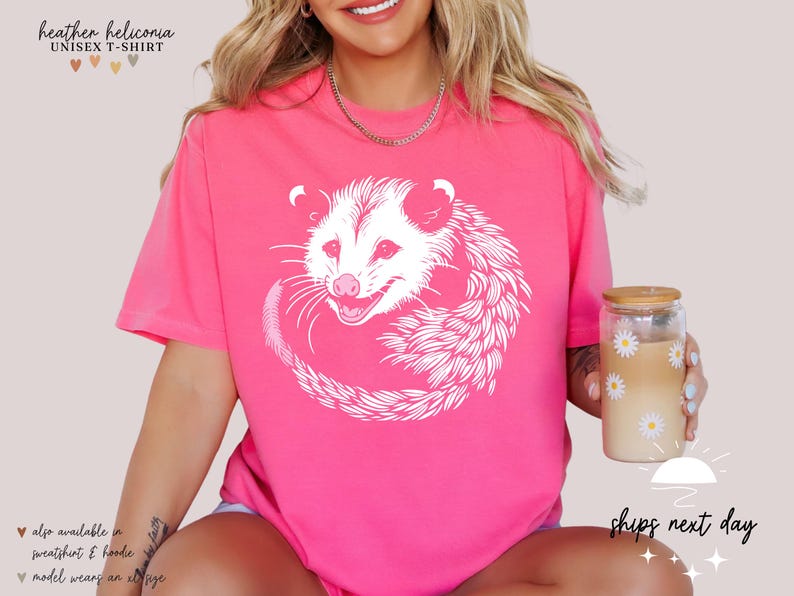 Puede incluir: Camiseta unisex rosa brillante con un dise&ntilde;o de zarig&uuml;eya blanca. La zarig&uuml;eya est&aacute; enroscada. El texto "heather heliconia UNISEX T-SHIRT" est&aacute; en la parte superior. Tambi&eacute;n disponible en sudadera y sudadera con capucha.