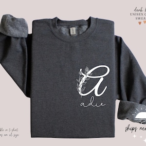 May include: Dark heather grey unisex crewneck sweatshirt with a white floral design and the name "adie" printed on the front.