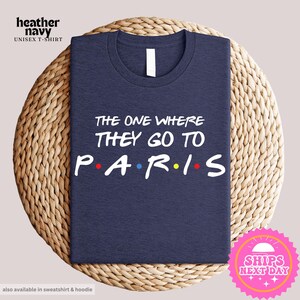 May include: A navy blue t-shirt with the text "THE ONE WHERE THEY GO TO PARIS" in white. The "P", "A", "R", and "I" in "PARIS" are each decorated with a small colored dot, reminiscent of the iconic "Friends" logo.