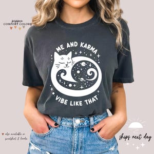 K&ouml;nnte beinhalten: Ein dunkelgraues T-Shirt mit einem wei&szlig;en Motiv eines Katers, der sich in einem Yin-Yang-Symbol mit Sternen und Planeten zusammengerollt hat. Der Text "Me and Karma Vibe Like That" ist unter dem Motiv aufgedruckt.