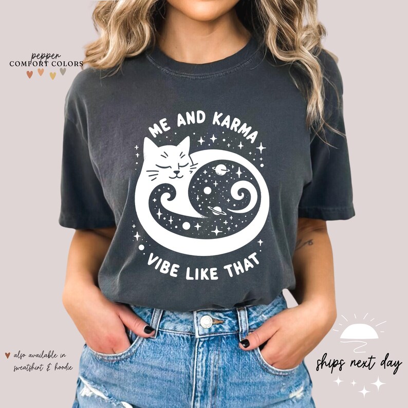K&ouml;nnte beinhalten: Ein dunkelgraues T-Shirt mit einem wei&szlig;en Motiv eines Katers, der sich in einem Yin-Yang-Symbol mit Sternen und Planeten zusammengerollt hat. Der Text "Me and Karma Vibe Like That" ist unter dem Motiv aufgedruckt.