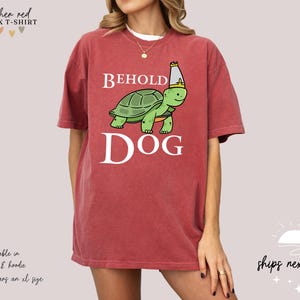 May include: A heather red unisex t-shirt featuring a cartoon turtle wearing a crown and the text "BEHOLD DOG".
