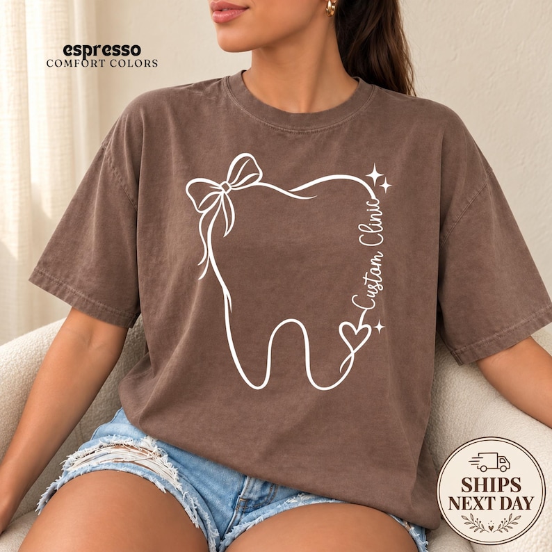 May include: An espresso-colored short-sleeve t-shirt featuring a white line-art graphic of a tooth adorned with a bow, a heart, and two small stars. The text "Custom Clinic" is printed vertically along the right side of the tooth design.