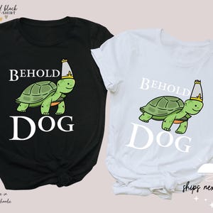 May include: Two t-shirts featuring a graphic of a turtle wearing a crown and the text "Behold Dog". One shirt is black and the other is white.