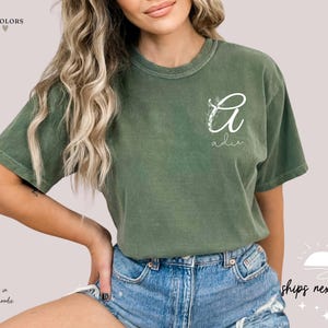 May include: Olive green t-shirt with a white floral design and the name "Adie" in script. The shirt is a comfortable fit and is paired with denim shorts. The text "Comfort Colors" and "ships next day" are also visible.