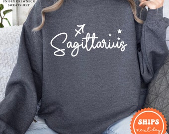 Könnte beinhalten: Dunkelgrauer Sweatshirt mit dem Text "Sagittarius" in weißer Kursivschrift und einem weißen Pfeilsymbol über dem Text. Rechts neben dem Text befinden sich drei weiße Sterne.