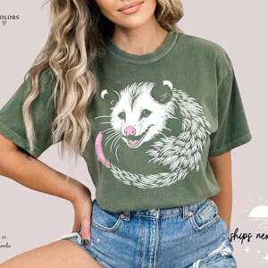 Puede incluir: Camiseta verde oliva con un dise&ntilde;o de zarig&uuml;eya blanca, nariz y cola rosas. La zarig&uuml;eya est&aacute; enroscada en un c&iacute;rculo. La parte superior de la camiseta tiene el texto "more comfort colors".