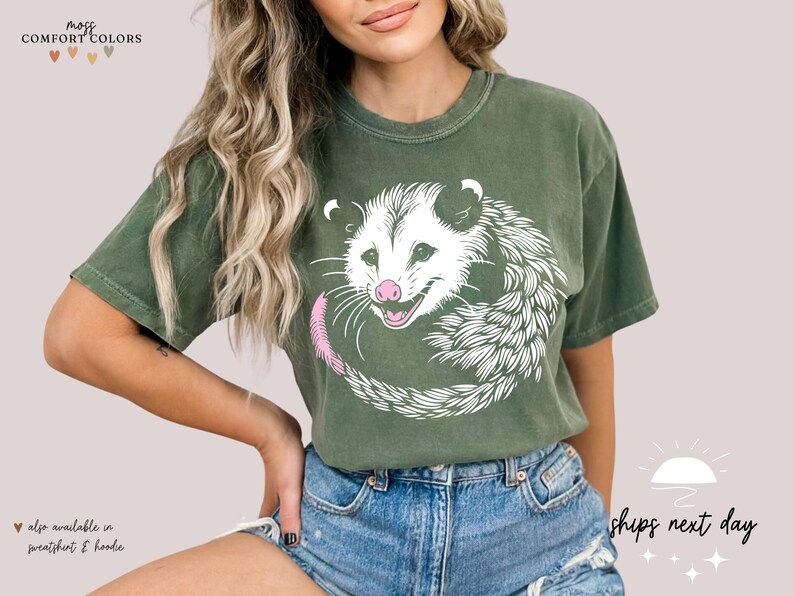 Puede incluir: Camiseta verde oliva con un dise&ntilde;o de zarig&uuml;eya blanca, nariz y cola rosas. La zarig&uuml;eya est&aacute; enroscada en un c&iacute;rculo. La parte superior de la camiseta tiene el texto "more comfort colors".