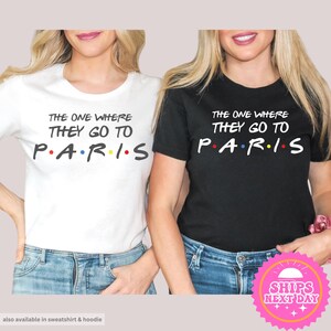 May include: Two white and black t-shirts with the text "The One Where They Go To Paris" with colorful dots representing the letters. The text is in a black font. The shirts are on two women wearing jeans.