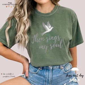 Puede incluir: Una camiseta verde con un p&aacute;jaro blanco y el texto "Then sings my soul" impreso en la parte delantera.