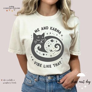 K&ouml;nnte beinhalten: Ein wei&szlig;es T-Shirt mit einem schwarzen Katzendesign und dem Text "Me and Karma Vibe Like That".