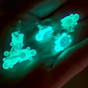 Puede incluir: Cuatro encantos que brillan en la oscuridad con forma de tortuga. Los encantos son blancos con un brillo azul claro.