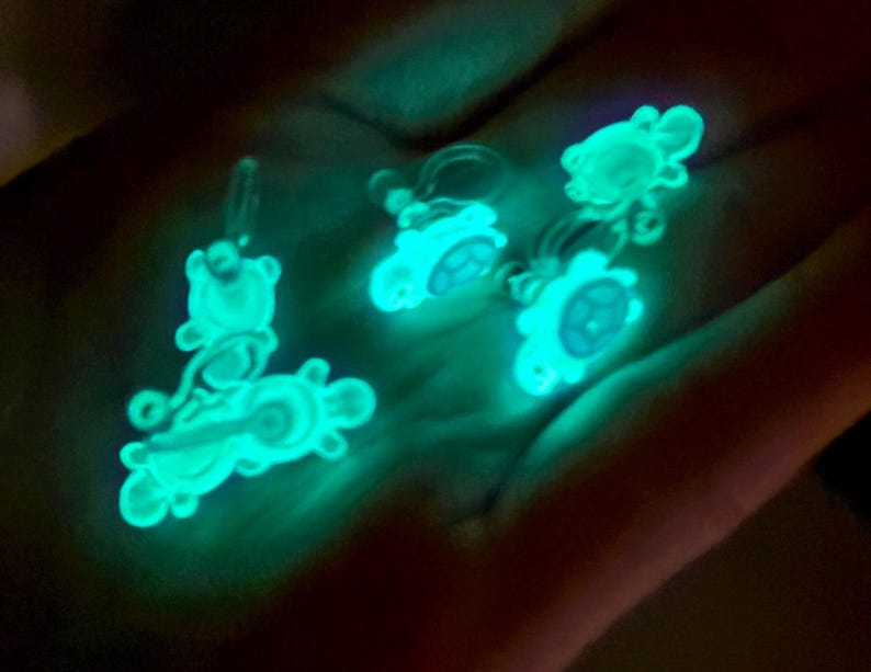 Puede incluir: Cuatro encantos que brillan en la oscuridad con forma de tortuga. Los encantos son blancos con un brillo azul claro.