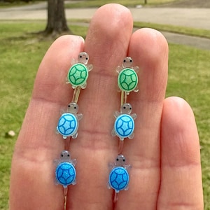 May include: Six small glass turtle charms, two green, two blue, and two light blue, with clear glass bodies and painted details. The charms are hanging from silver wires.