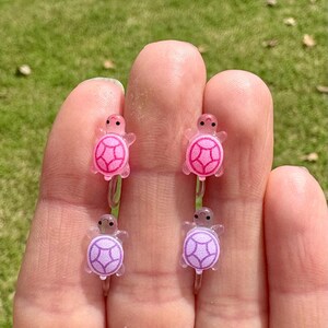 Puede incluir: Cuatro peque&ntilde;os amuletos de tortuga, dos rosas y dos morados, con un acabado de pl&aacute;stico transparente. Los amuletos est&aacute;n en un anillo de pl&aacute;stico transparente.