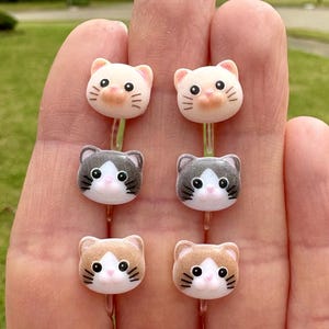 May include: Six pairs of cat-shaped hair clips. The clips are made of plastic and have a white base with different colored accents. The cats have black eyes, pink noses, and black whiskers.