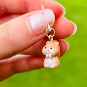 May include: A miniature hamster earring charm. The small, detailed figurine is tan and white, with black eyes and a pink nose. The earring is held by a silver-colored metal loop and a clear plastic post.