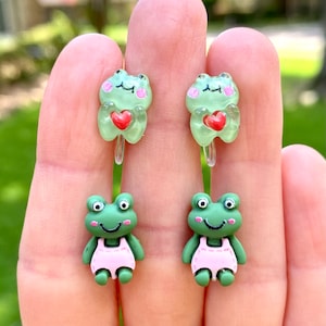 May include: A pair of green frog earrings. One frog is holding a red heart and the other frog is wearing a pink swimsuit.