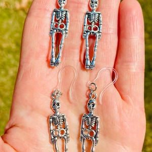 May include: A pair of silver skeleton earrings with clear plastic hooks. The skeletons are standing upright with their arms at their sides.